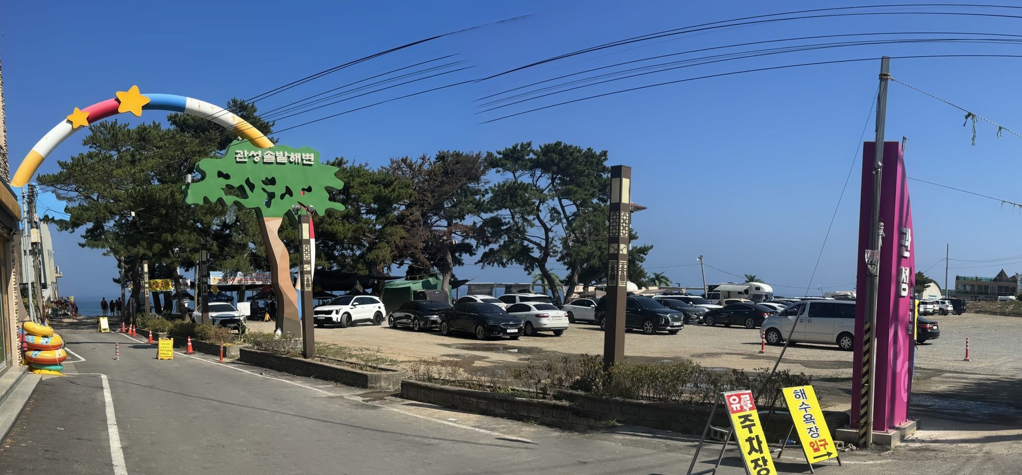 관성솔밭해변 입구 — 무지개 아치와 소나무 모양 간판, 옆에 무료 주차장